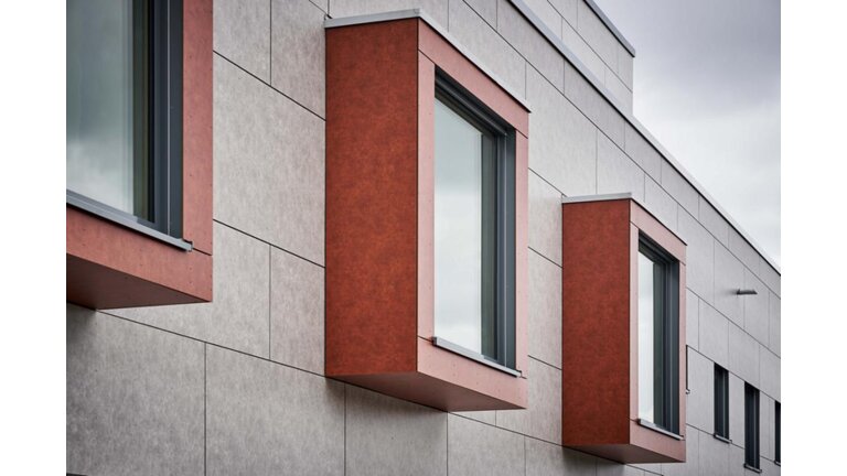 Moderne Fassade mit hervorstehenden, rotbraunen Fensterrahmen und grauen Paneelen, aufgenommen an einem bewölkten Tag.